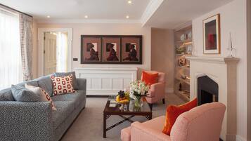 A living room with a sofa, armchair, coffee table, and fireplace. There are framed pictures on the wall and a vase with flowers on the coffee table.