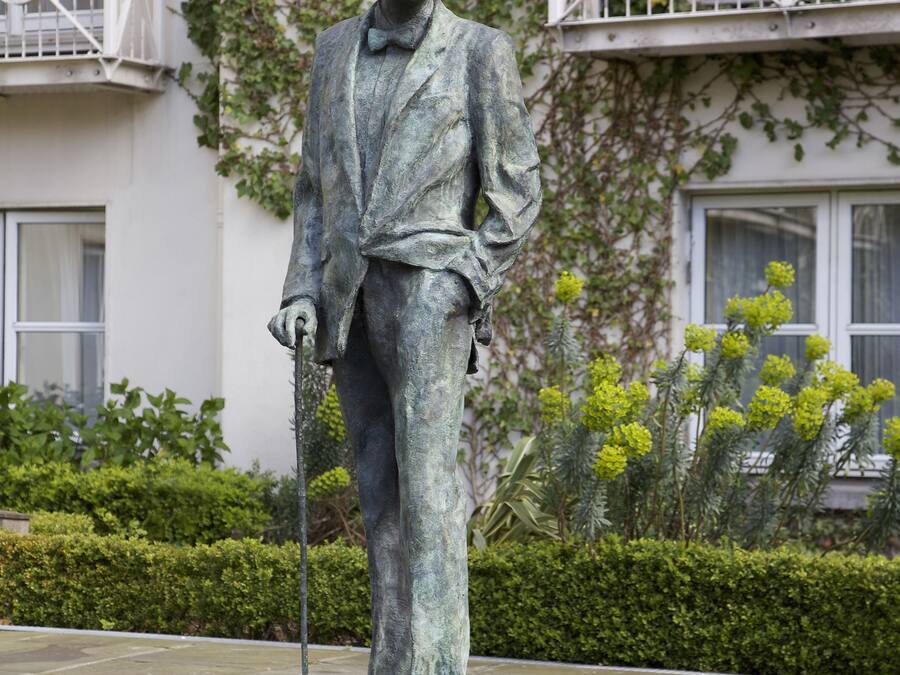 Una estatua de un hombre con traje y sombrero, de pie sobre una plataforma circular en un patio con un edificio al fondo.