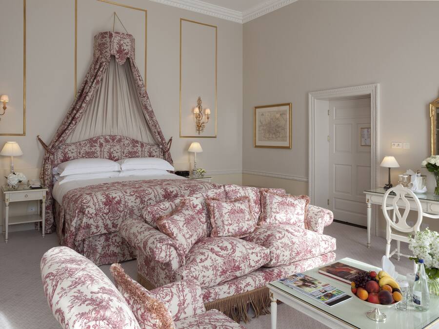 A bedroom with a canopy bed, a matching bench, a bedside table with a lamp, a mirror, and a floral arrangement on a side table.