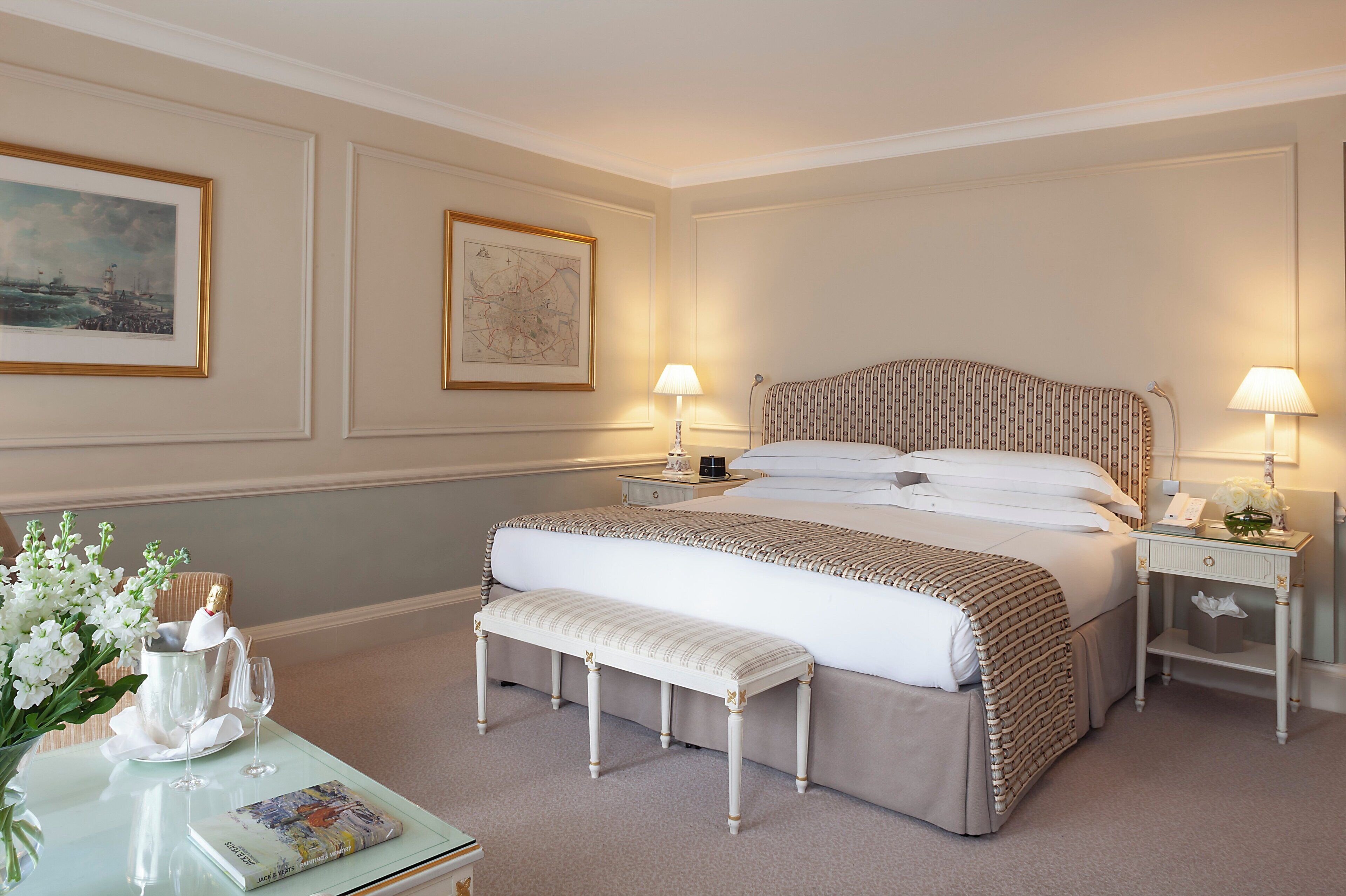 Een keurig opgemaakt slaapkamer met een groot bed, twee nachtkastjes, een bankje en een tafeltje met een bloemenvaas.
