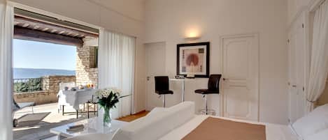 A bedroom with a bed, a sitting area with a table and chairs, a large window with a view, and a door.