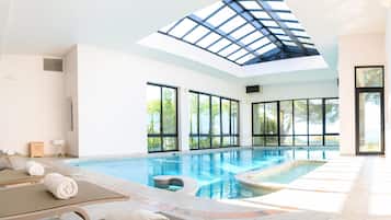 Une grande piscine intérieure avec une verrière, de nombreuses fenêtres et des chaises longues avec des serviettes.