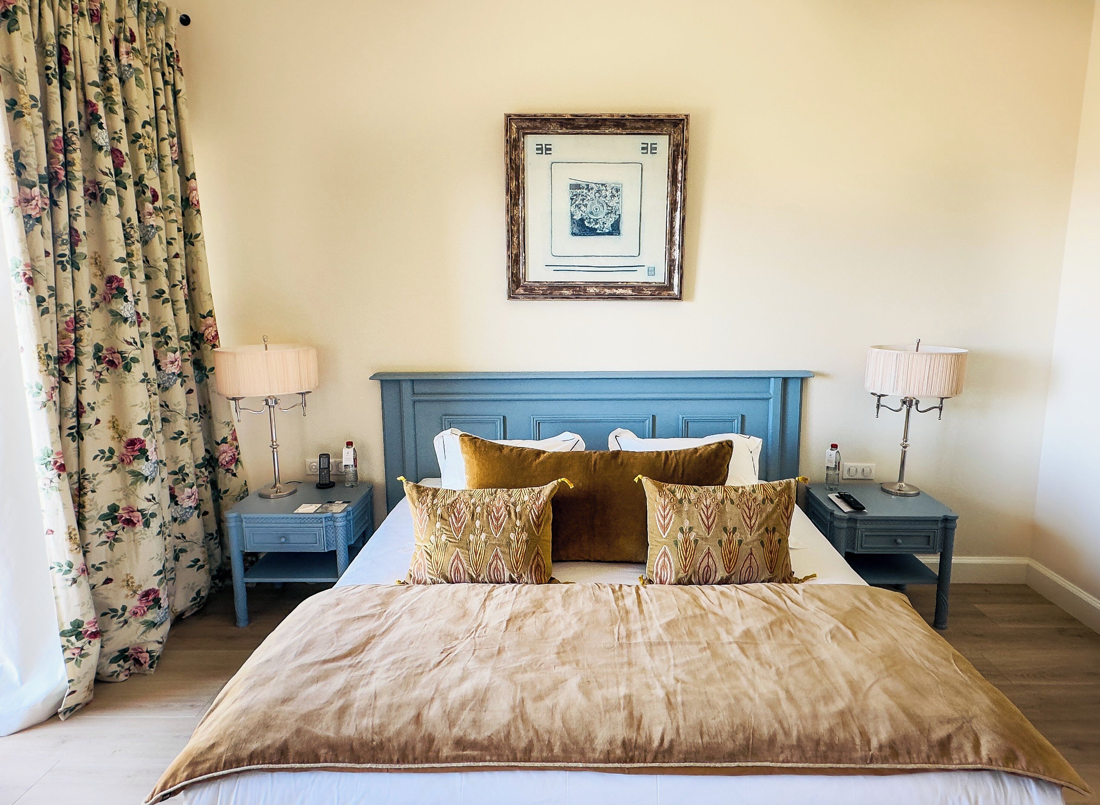 A neatly arranged bedroom with a large bed, floral curtains, a blue headboard, and two bedside tables with lamps and a bottle.