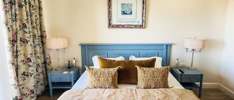 A neatly arranged bedroom with a large bed, floral curtains, a blue headboard, and two bedside tables with lamps and a bottle.