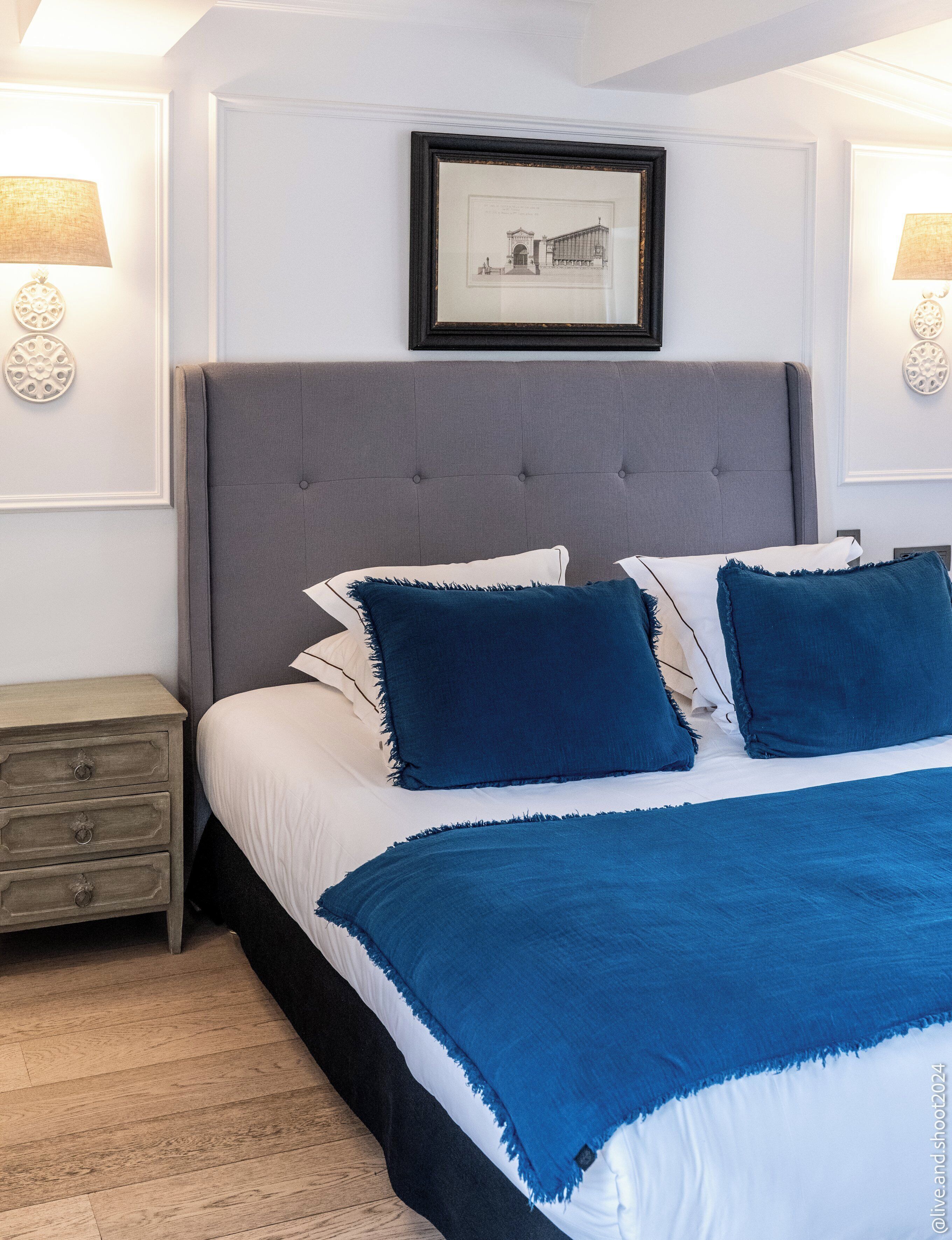 Une chambre avec une tête de lit rembourrée de couleur grise, une literie blanche et un plaid bleu. Un tableau encadré est accroché au mur au-dessus du lit, et une petite table de chevet avec une lampe se trouve à côté du lit.