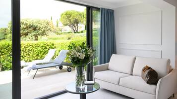 A modern living room with a white sofa, a glass table with a vase of flowers, and a view of a garden through large windows.
