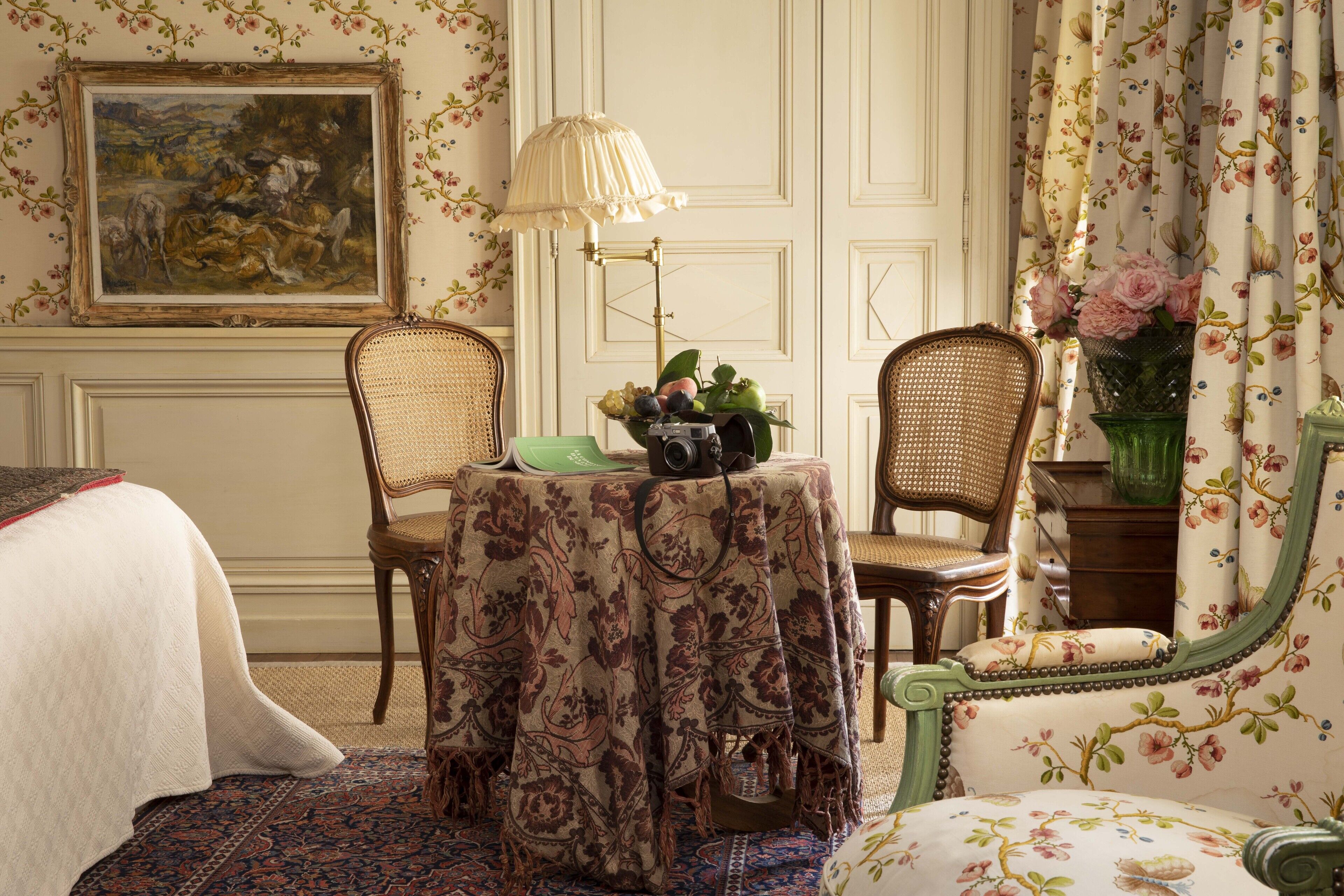 A hotel room with a bed, two chairs, a table with a floral tablecloth, a lamp, and a painting on the wall.