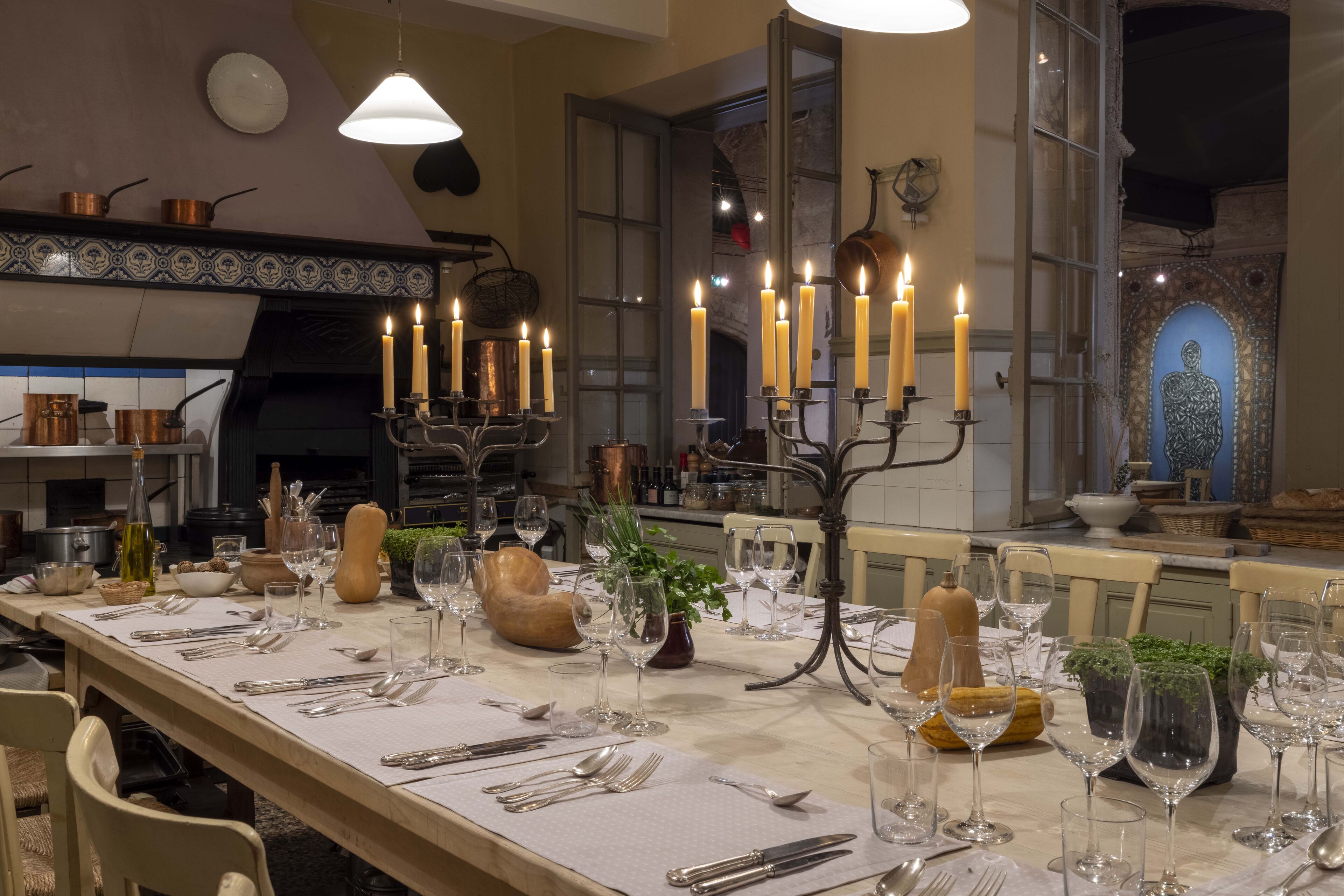 Una mesa de comedor dispuesta con velas, copas de vino y cubiertos, rodeada de sillas y utensilios de cocina.