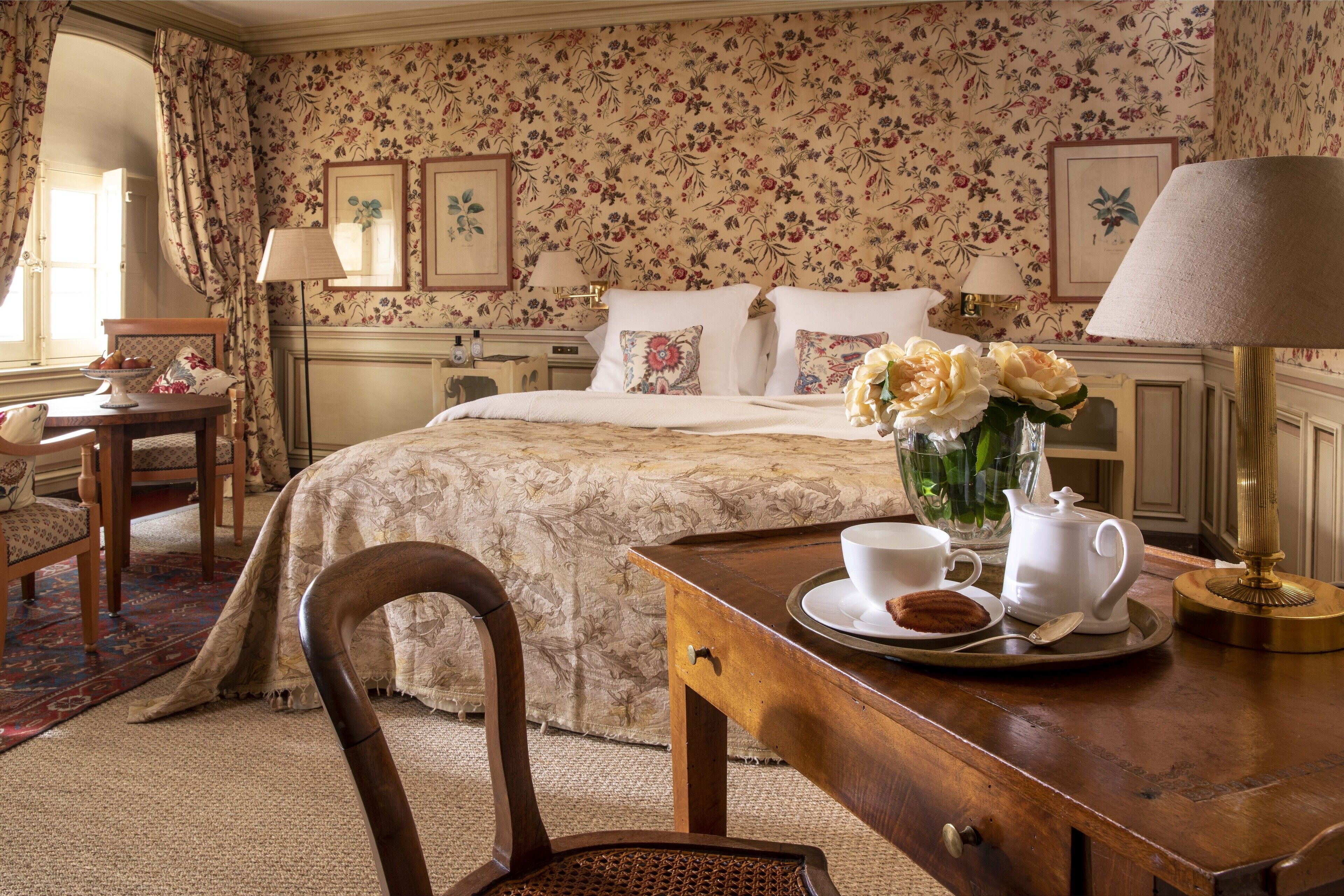 A neatly arranged bedroom with floral wallpaper, a bed with white linens, a bedside table with a lamp, a chair, and a tray with a teapot and cups.