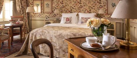 A neatly arranged bedroom with floral wallpaper, a bed with white linens, a bedside table with a lamp, a chair, and a tray with a teapot and cups.