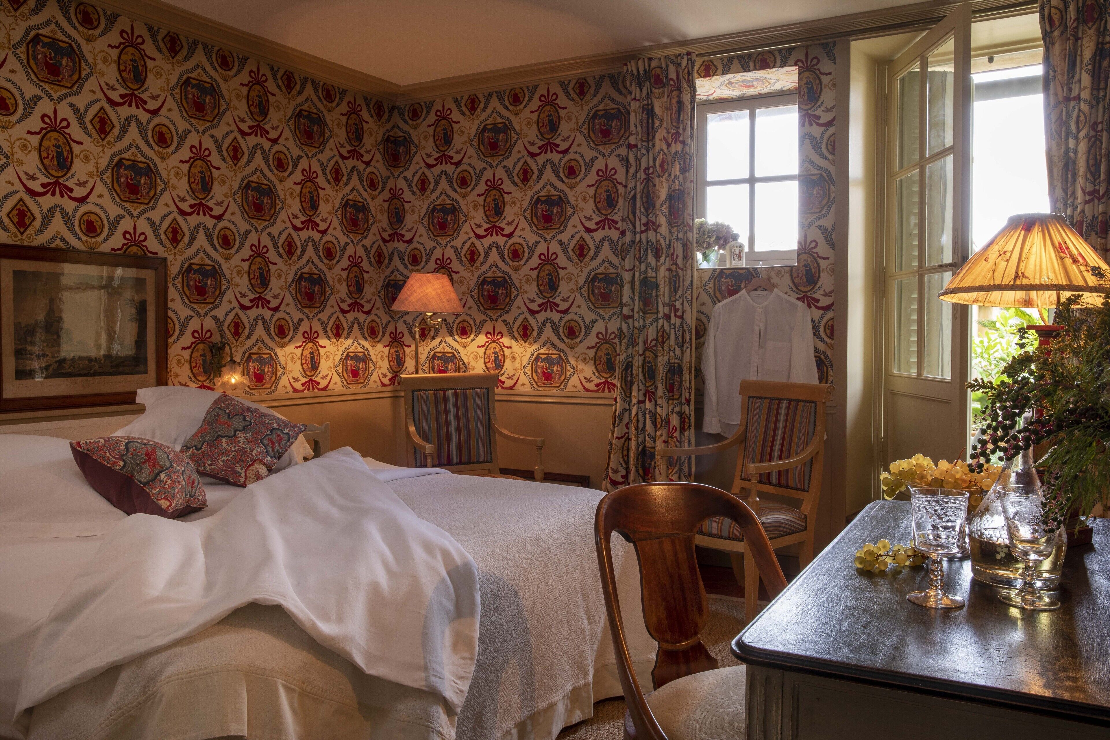 A hotel room with a bed, a wooden chair, a small table with a lamp, and a window with curtains.