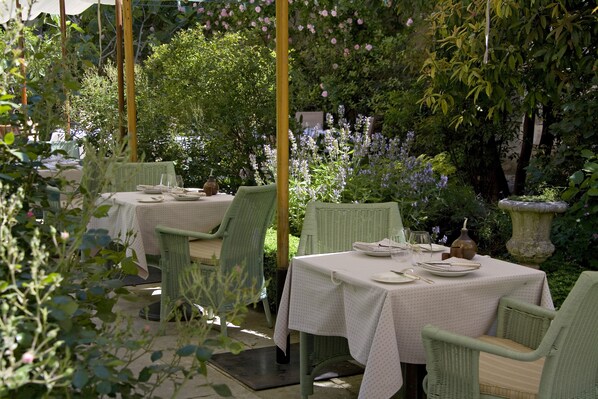 An outdoor dining area with tables and chairs, surrounded by lush greenery and flowering plants.