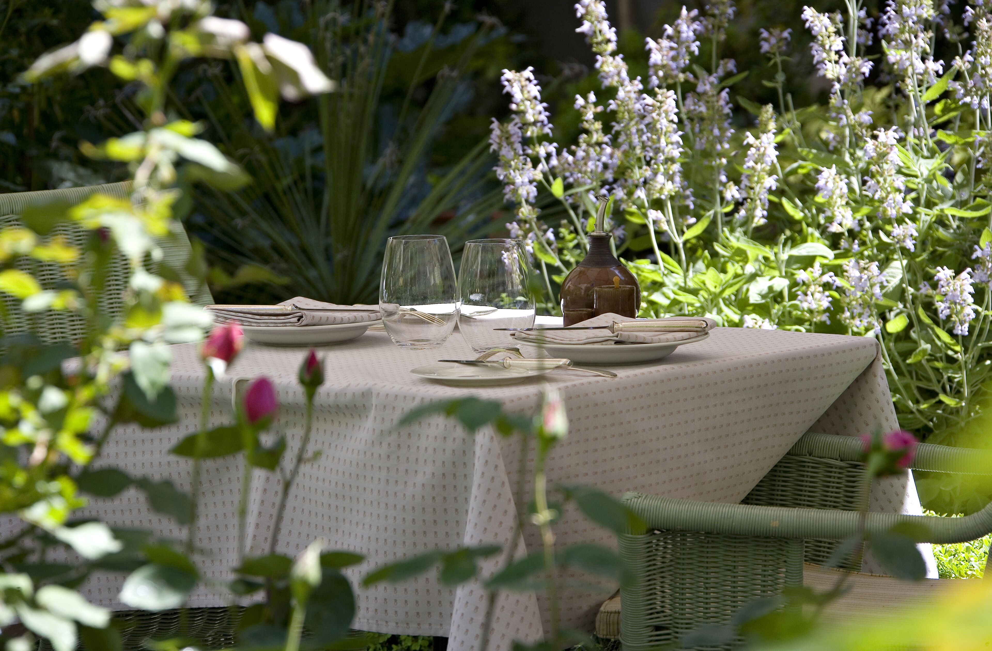 A table set outdoors with a white tablecloth, surrounded by greenery and flowers.