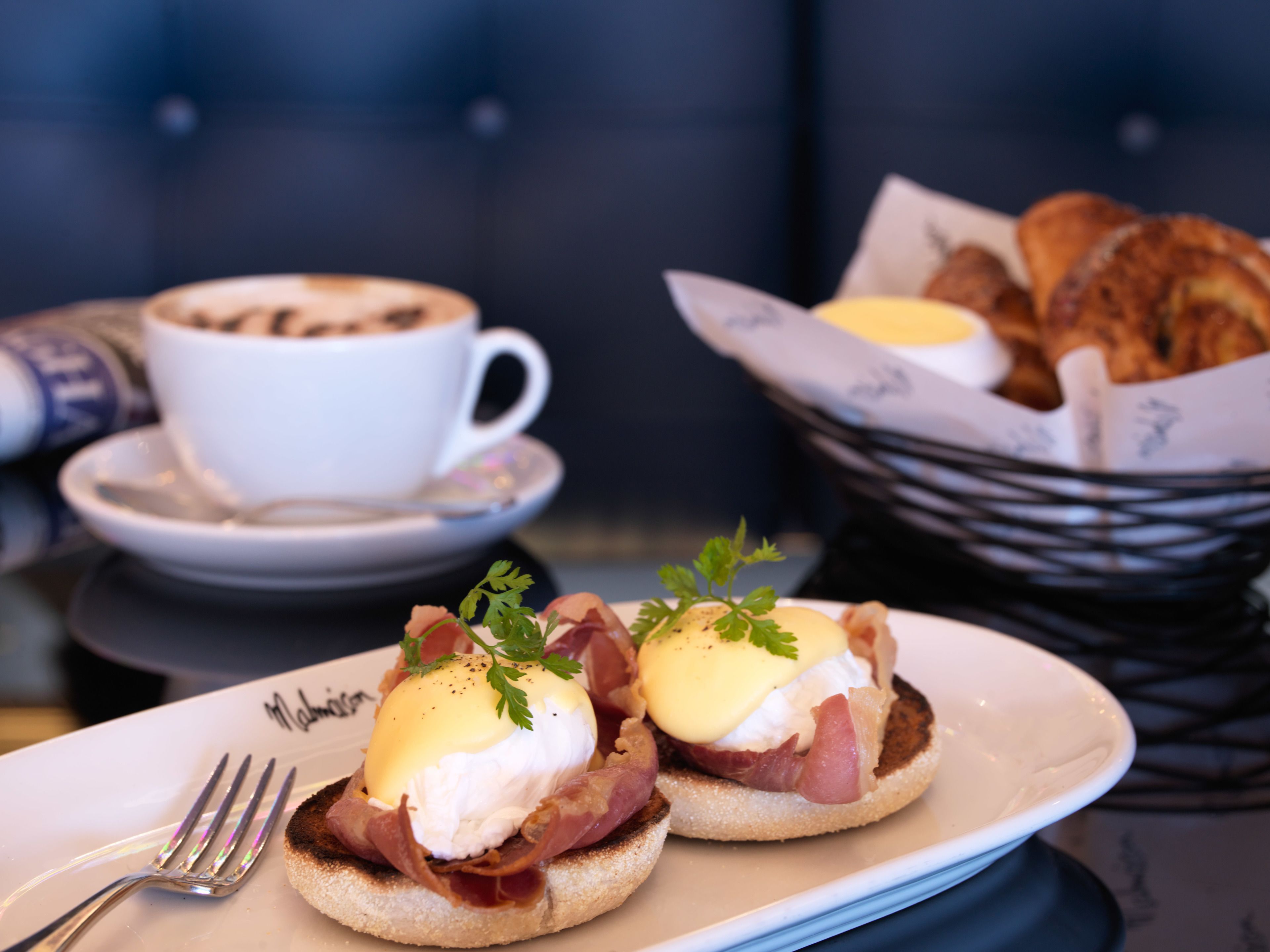 A breakfast plate with eggs benedict, a cup of coffee, and a basket of croissants.