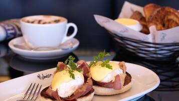 A breakfast plate with eggs benedict, a cup of coffee, and a basket of croissants.