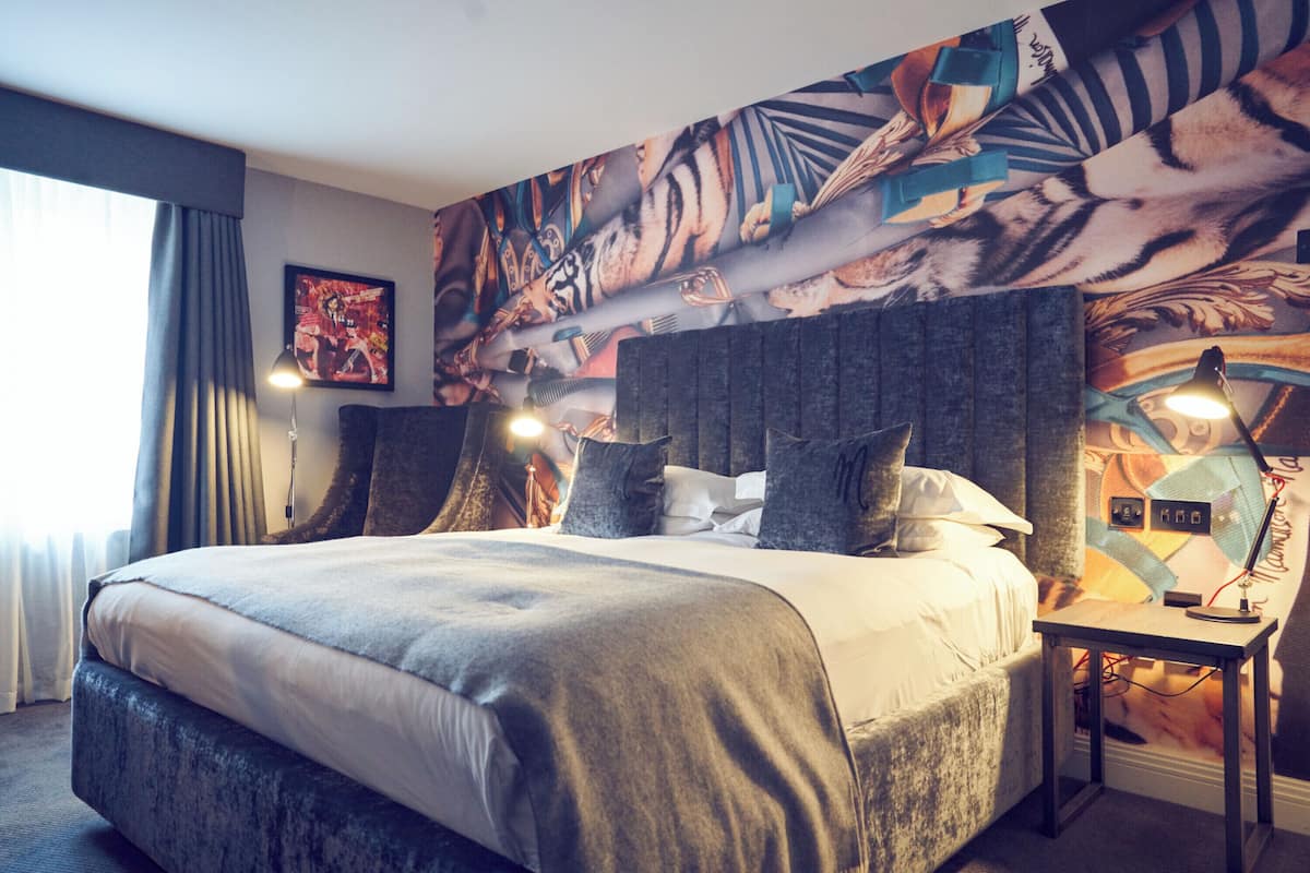 A large bed with a grey headboard, two bedside tables with lamps, a wall with a tiger and abstract art, and a window with curtains.