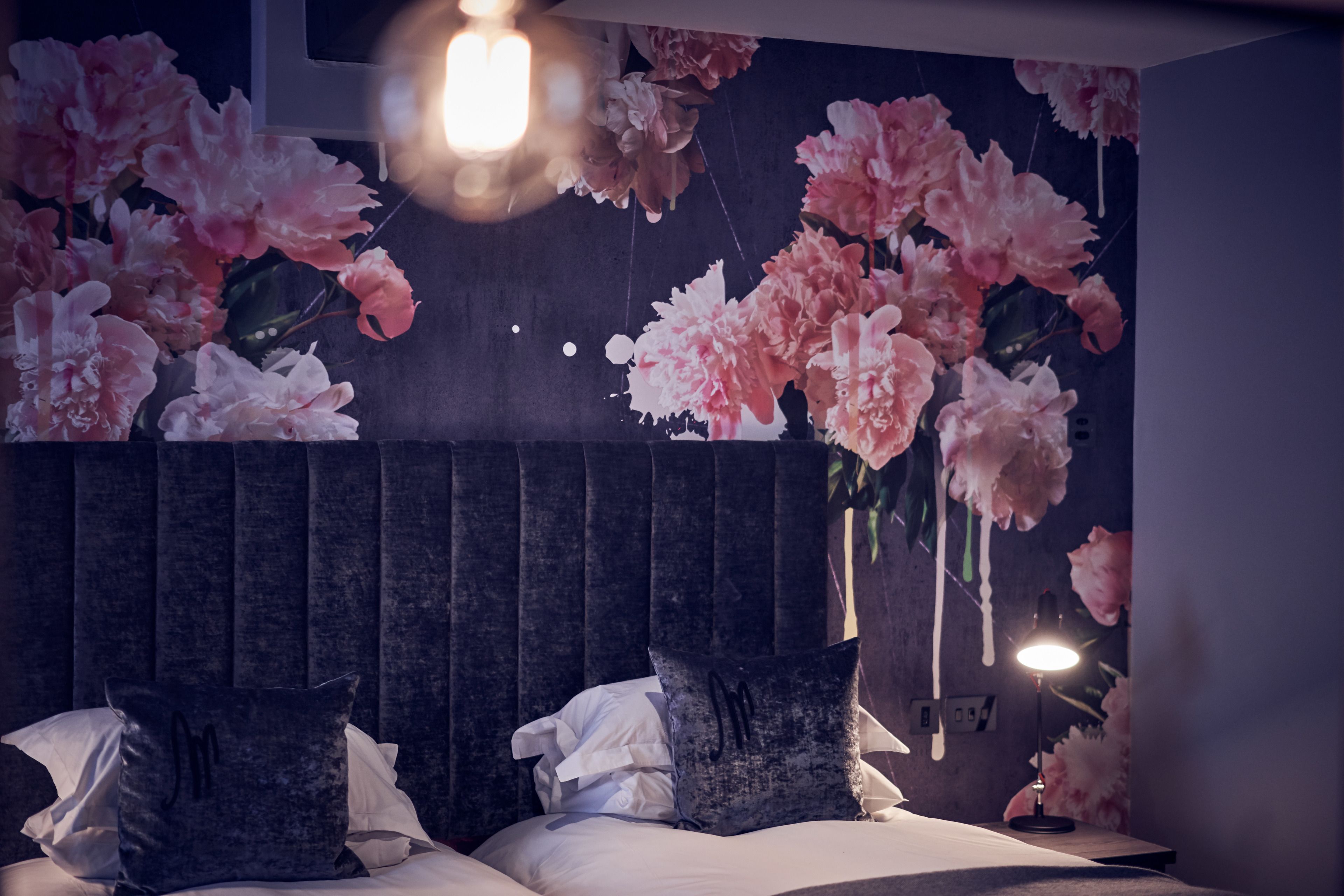 Quarto com cama, travesseiros, mesa de cabeceira, luminária na parede e um mural floral.
