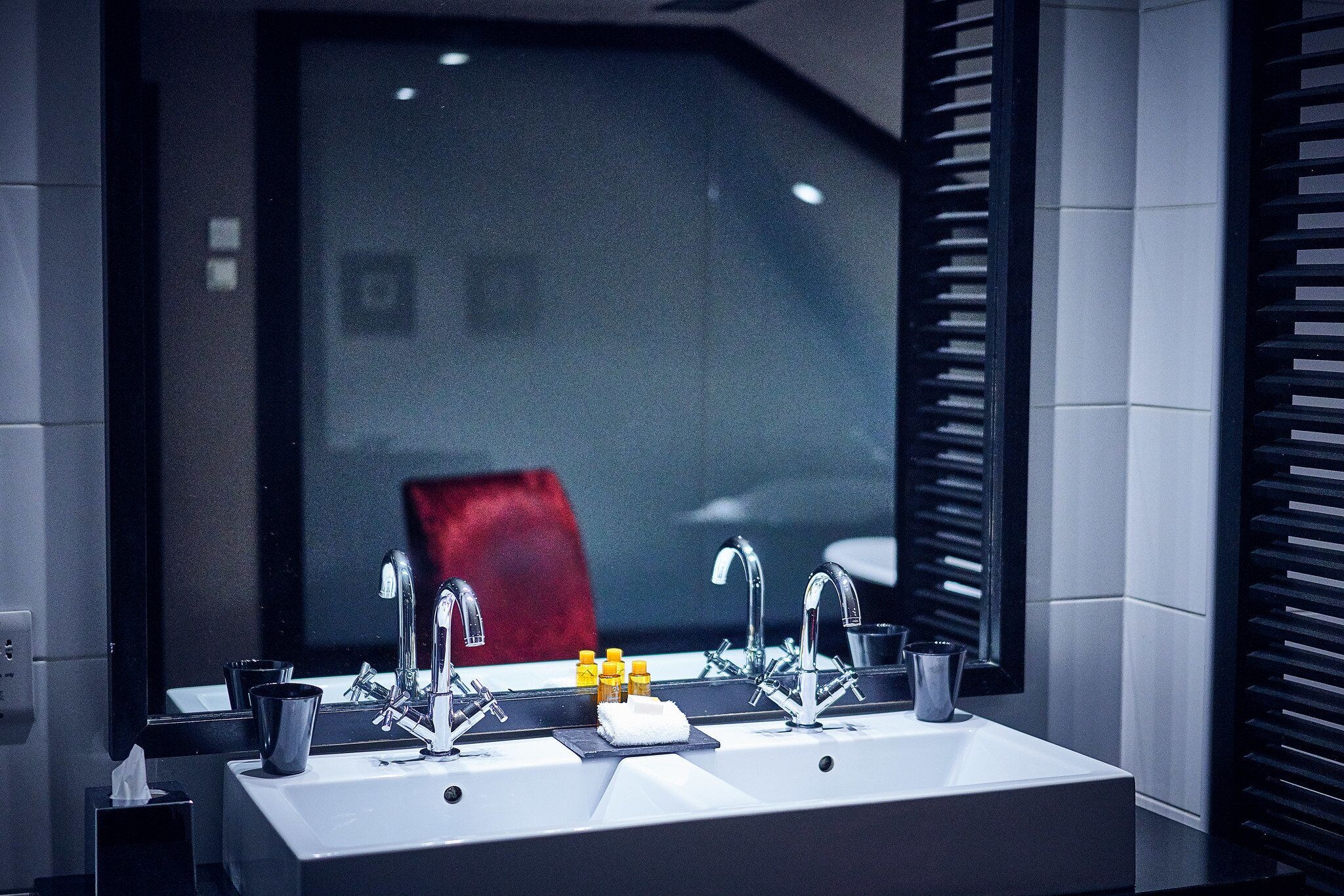 A modern bathroom with a large mirror, two sinks, and a red chair in the background.
