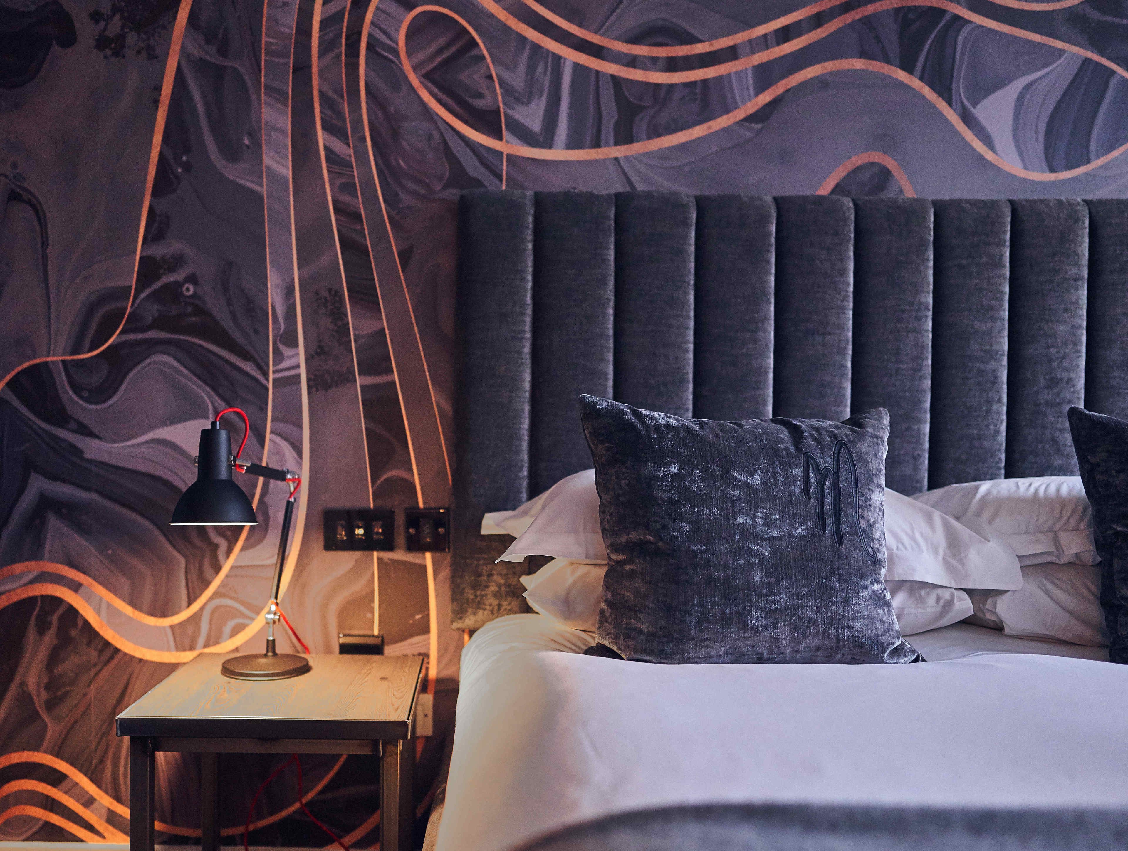 A modern bedroom with a dark headboard, a bedside table with a lamp, and a patterned wall.