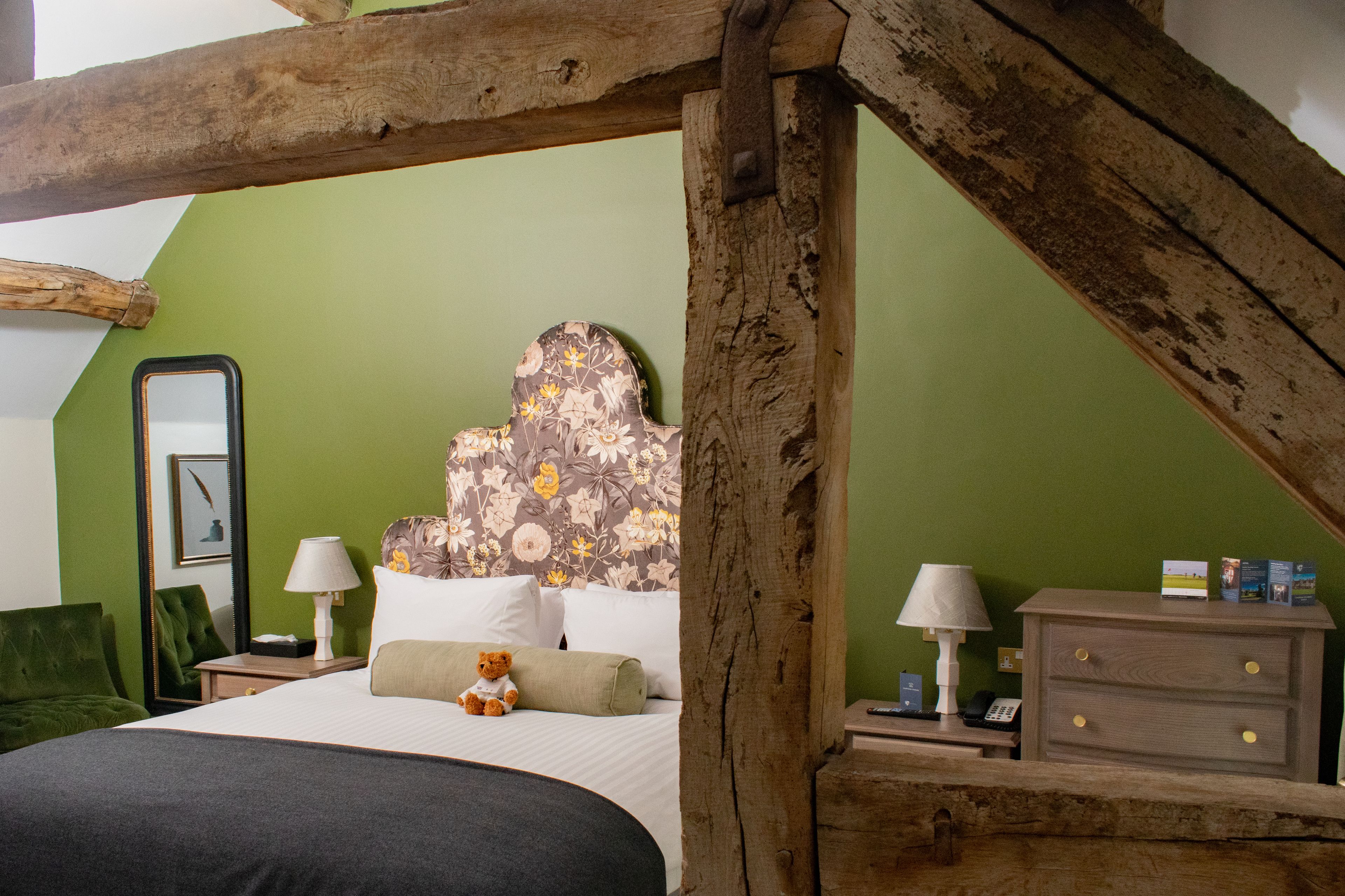 Un dormitorio con techo de madera, cama con ropa blanca, pared verde y cabecera con estampado floral.