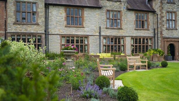 A stone building with multiple windows and a well-maintained garden with benches and flowers.