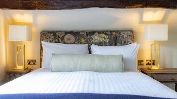 A neatly made bed with white linens and a blue throw blanket, flanked by two bedside tables with lamps, a floral headboard, and a wooden beam above.