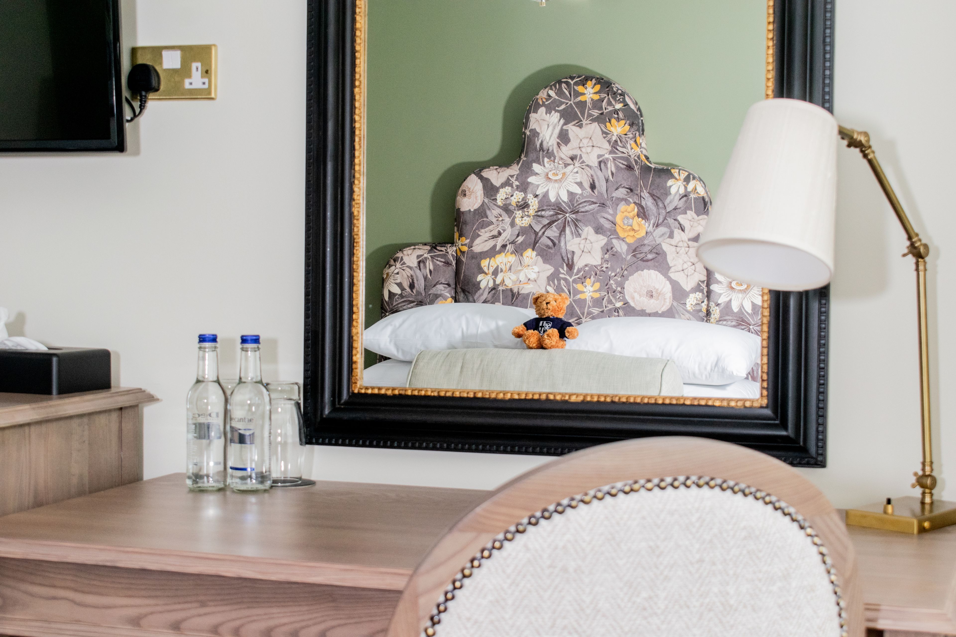 A hotel room with a bed, a bedside table with bottles, a lamp, and a mirror reflecting the bed.