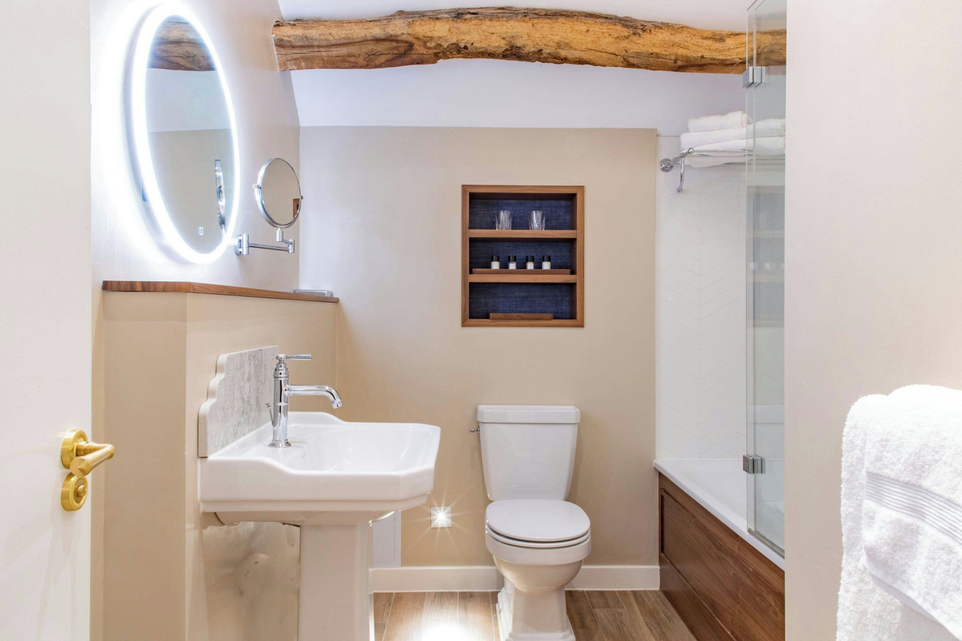 A bathroom with a white sink, a toilet, a wooden shelf with toiletries, and a glass shower enclosure.