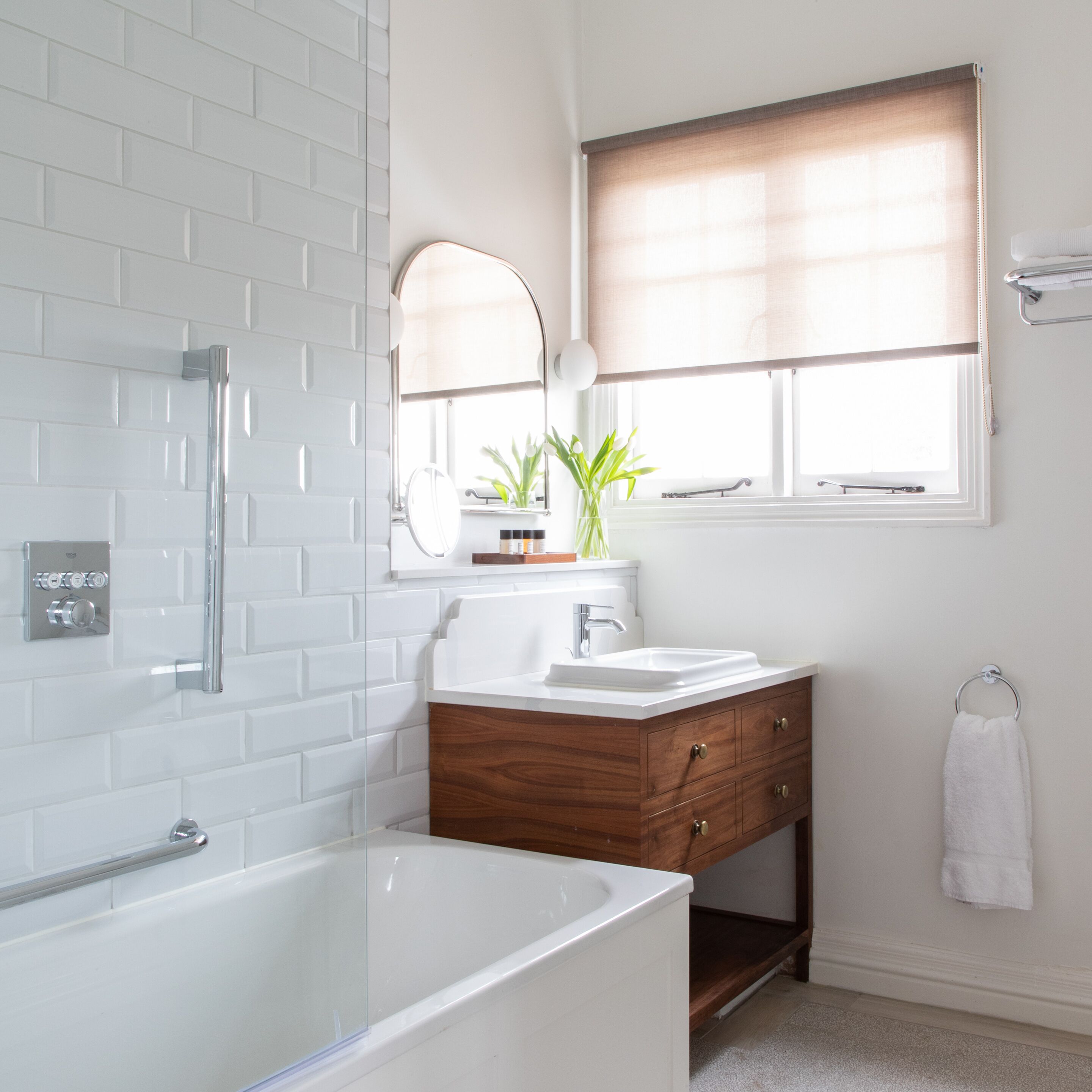 Een moderne badkamer met een witte badkuip, een houten wastafelmeubel, een spiegel en een raam met zonwering.