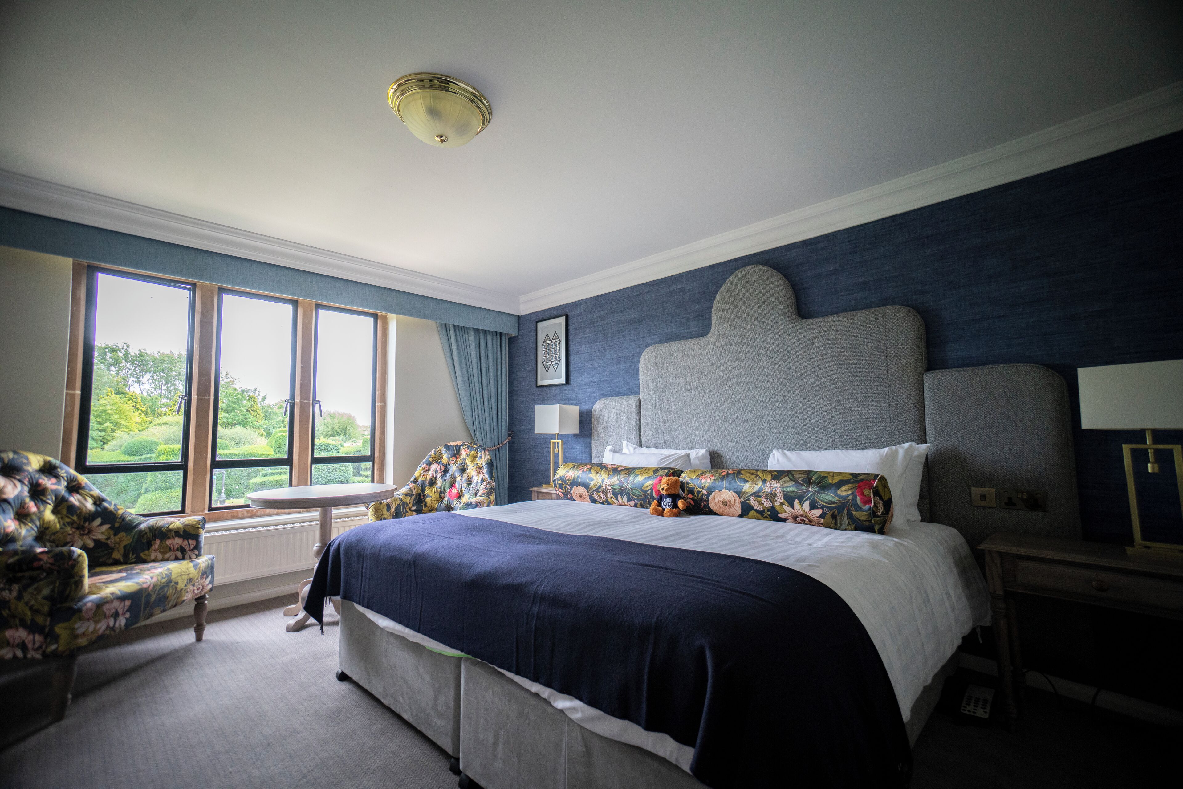 A hotel room with a large bed, a chair, a desk, and a window with a view of greenery.