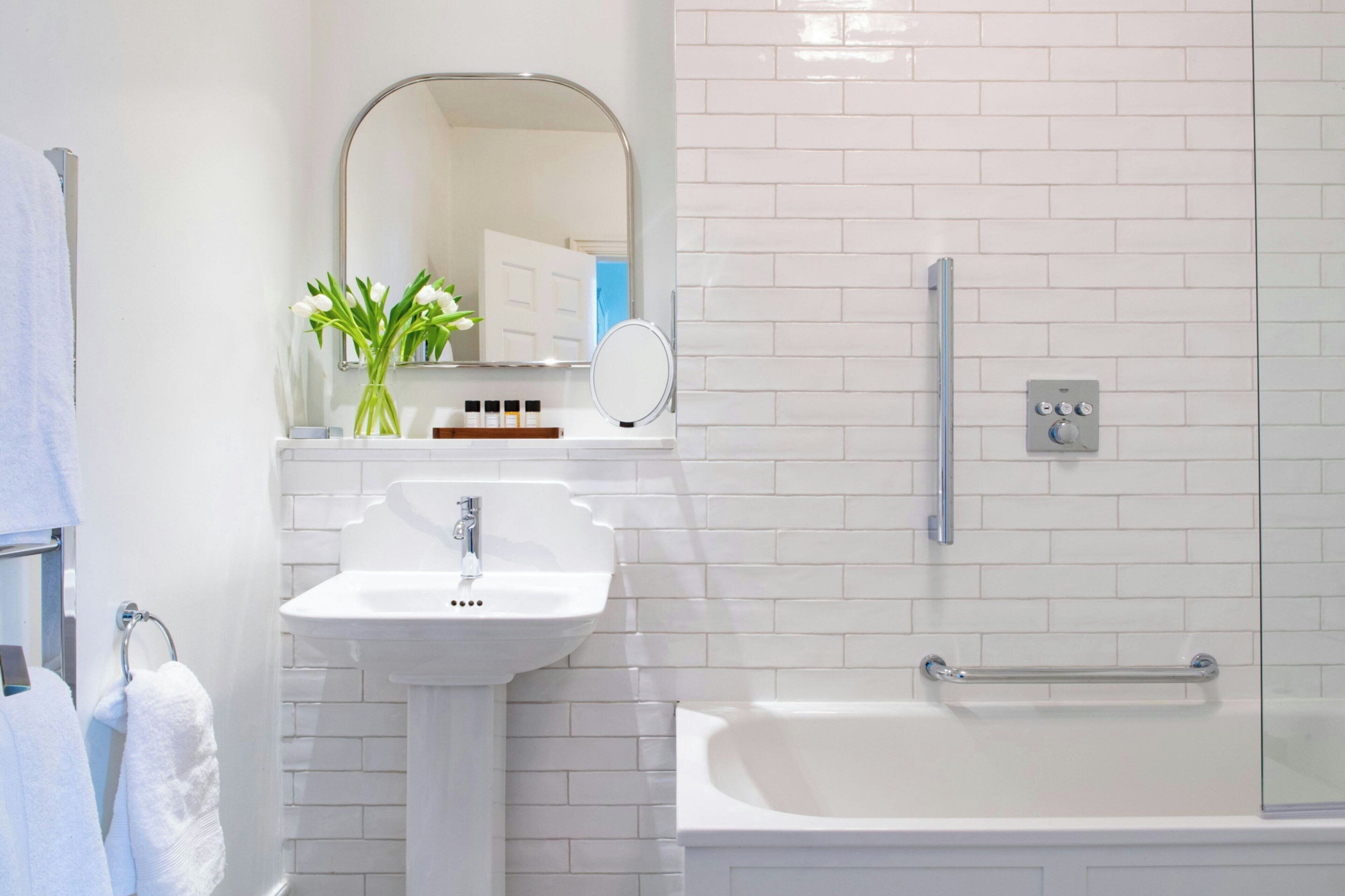 A bathroom with a white sink, a mirror, a bathtub with a grab bar, and a white tiled wall.