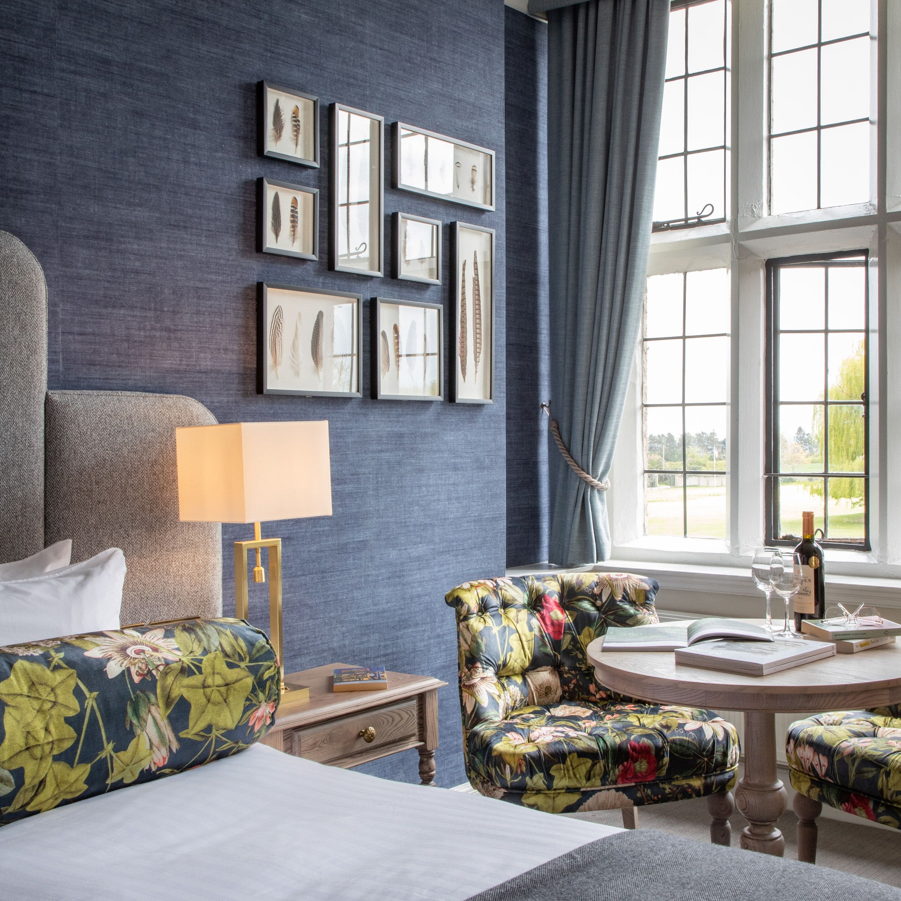 Een hotelkamer met een bed, een bloemen fauteuil, een ronde tafel met een wijnfles en glazen, en een wand met ingelijste foto's.