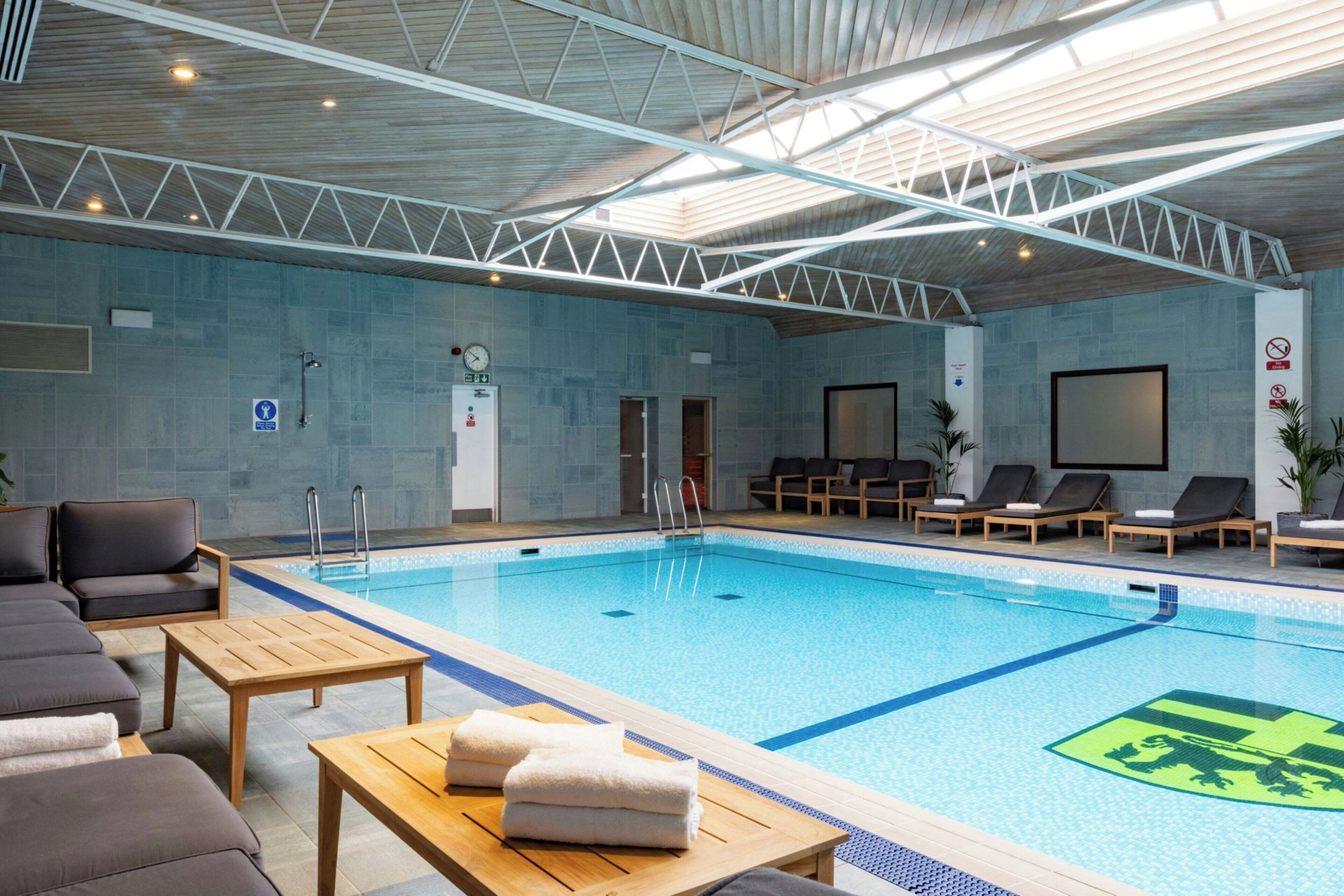 An indoor swimming pool with lounge chairs and a wooden table with towels.