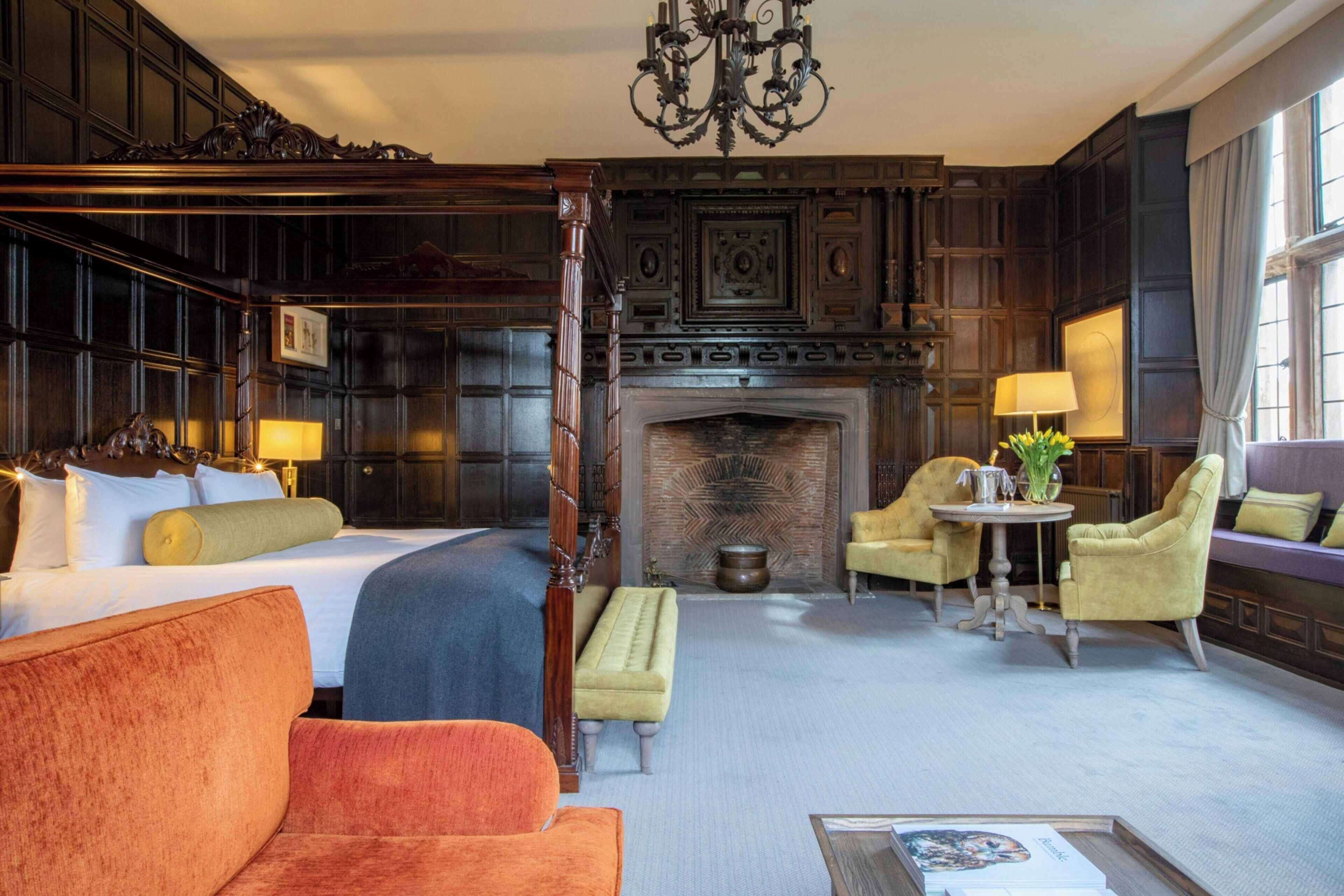 A hotel room with a large bed, a sitting area with a sofa and armchairs, a fireplace, and a dining table with lamps.