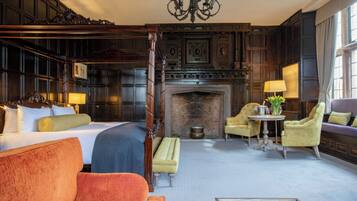 A hotel room with a large bed, a sitting area with a sofa and armchairs, a fireplace, and a dining table with lamps.