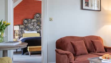 Een kamer met een bruine bank, een gele fauteuil, een tafeltje met eten erop en een bed met een bloemenmotief hoofdeinde op de achtergrond.