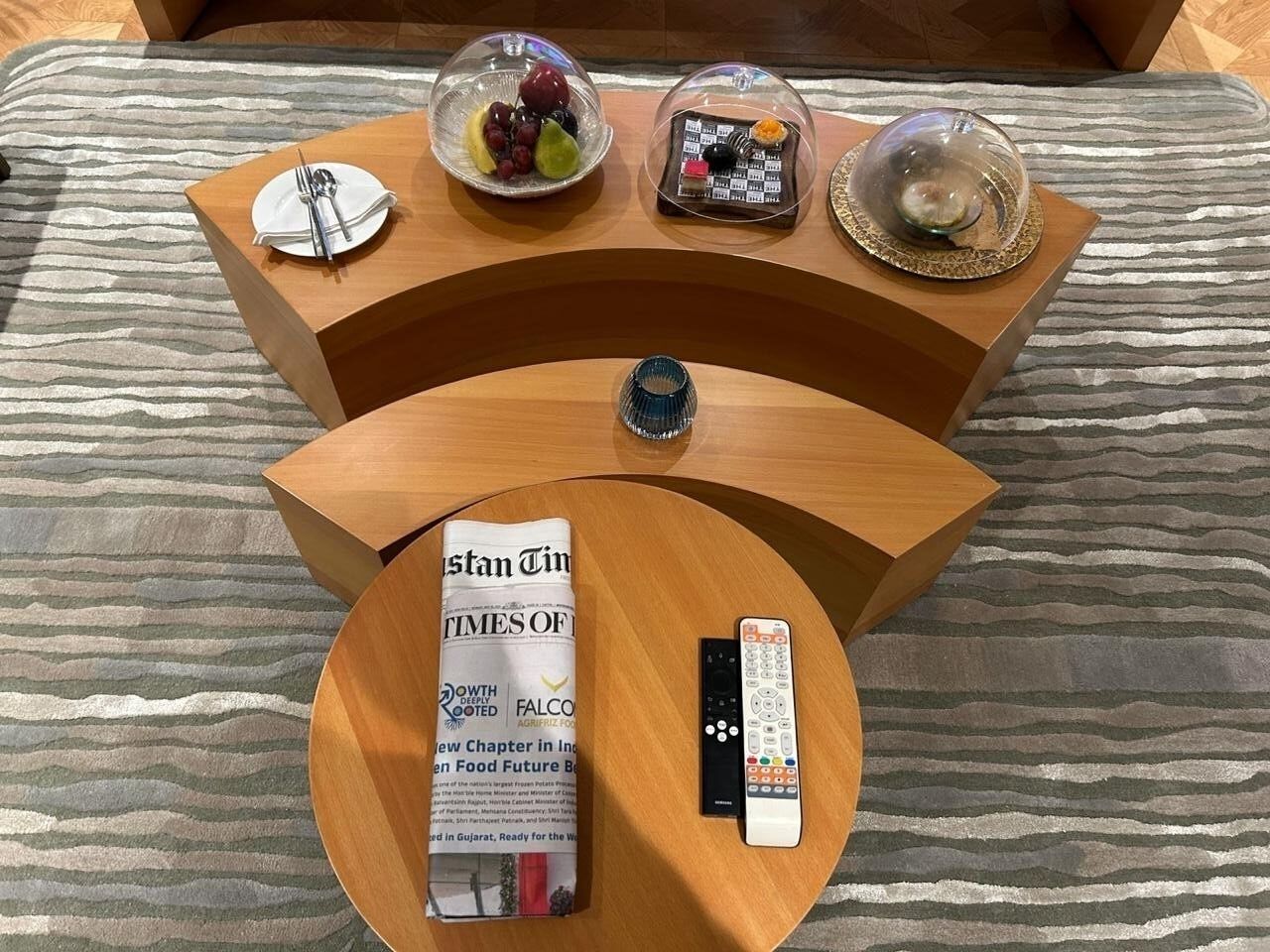 A hotel room with a wooden side table, a remote control, a book titled 'FALCON', a glass bowl with fruit, and a decorative item.
