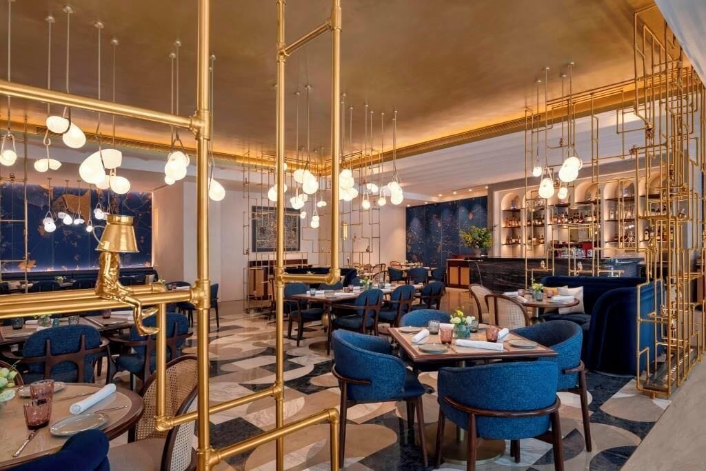 Un restaurante con mesas redondas, sillas azules, lámparas colgantes y una zona de bar.