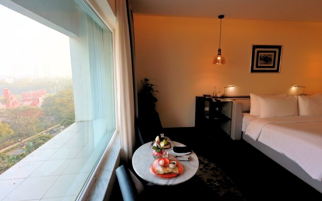 A hotel room with a bed, bedside table, lamp, and a view of a cityscape through a large window.