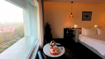 A hotel room with a bed, bedside table, lamp, and a view of a cityscape through a large window.