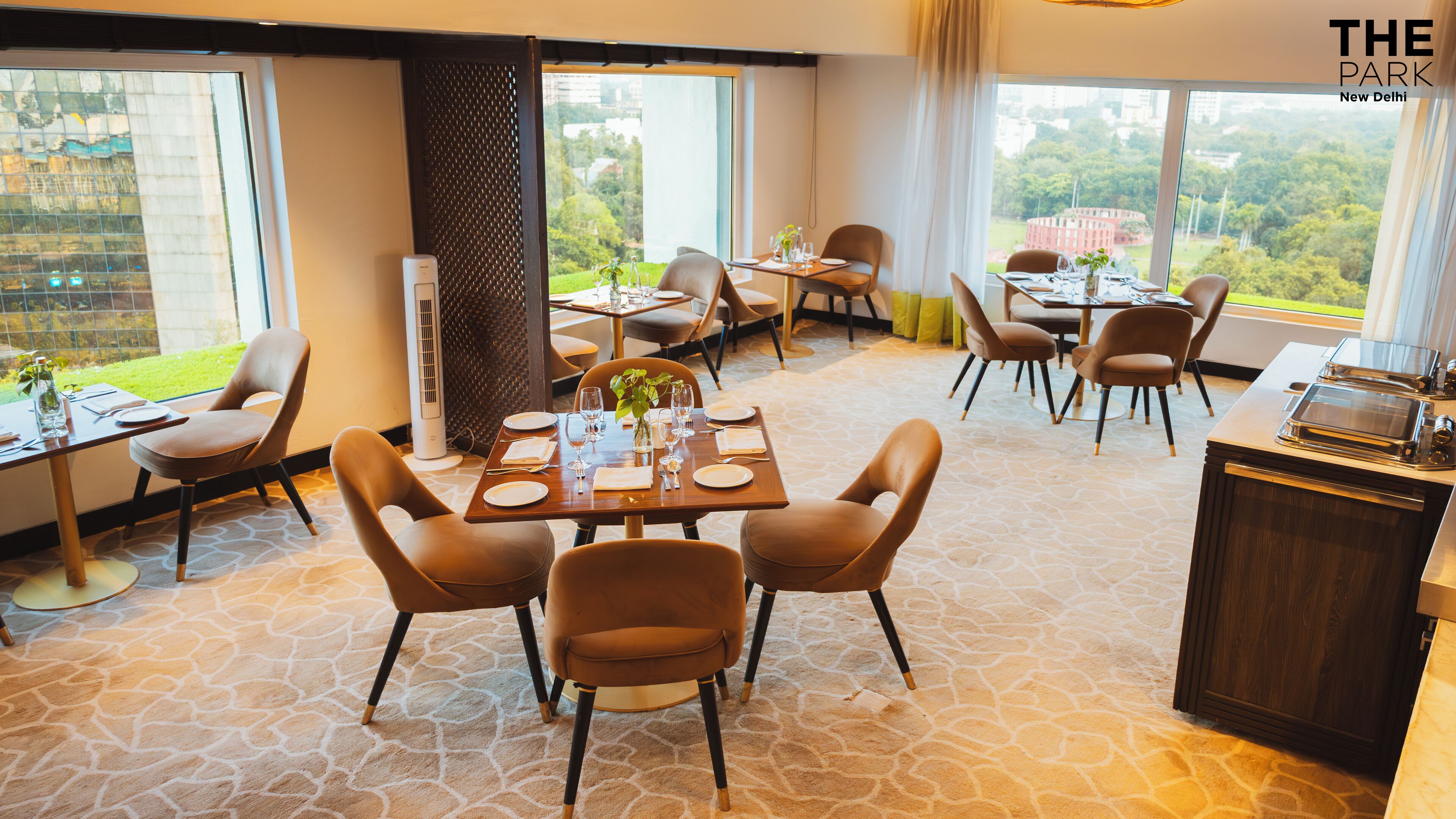A dining area with tables set for four, large windows offering a view of the city, and a modern kitchen area.