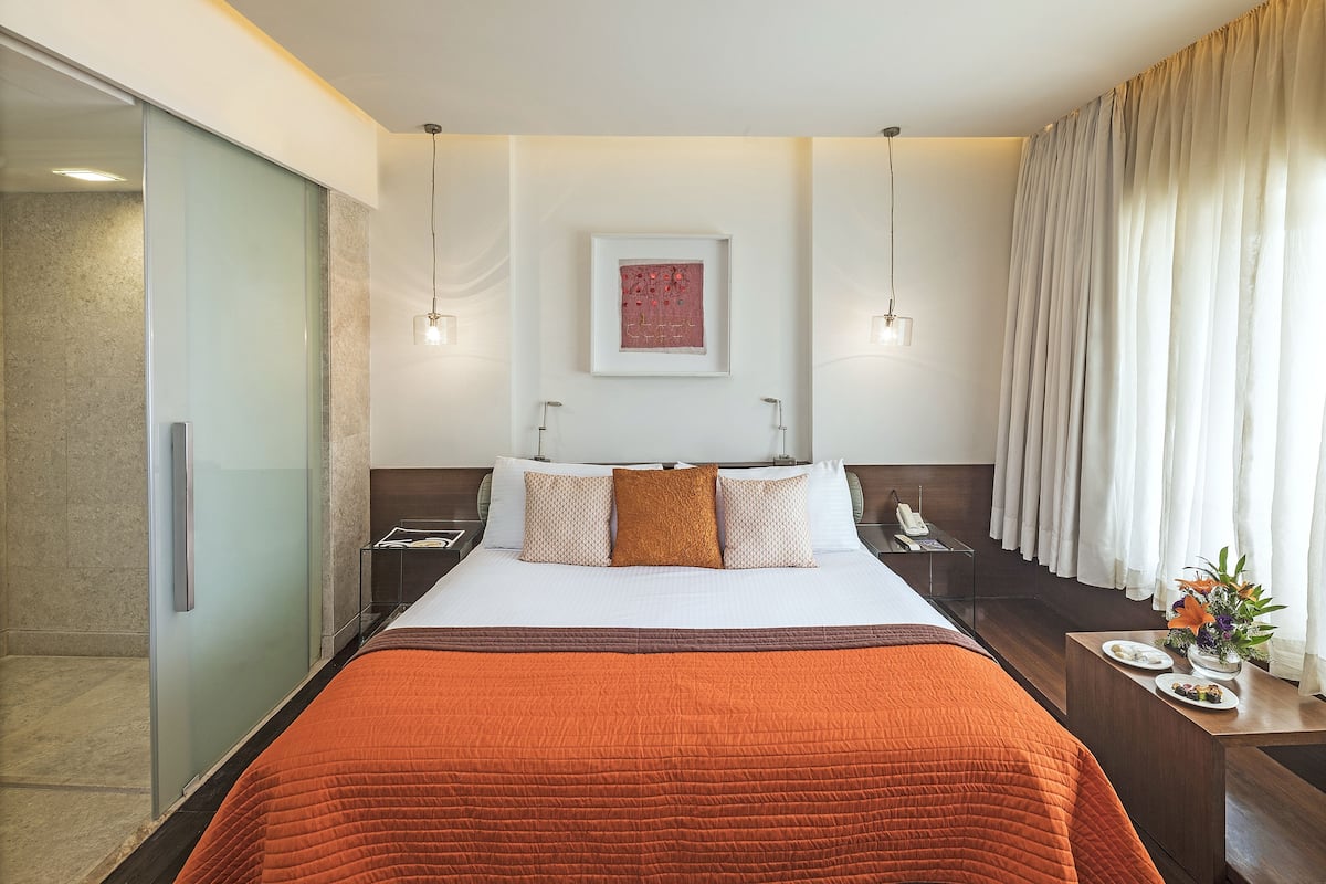 A hotel room with a large bed, a bedside table with a phone, a nightstand, a glass shower enclosure, and a window with curtains.