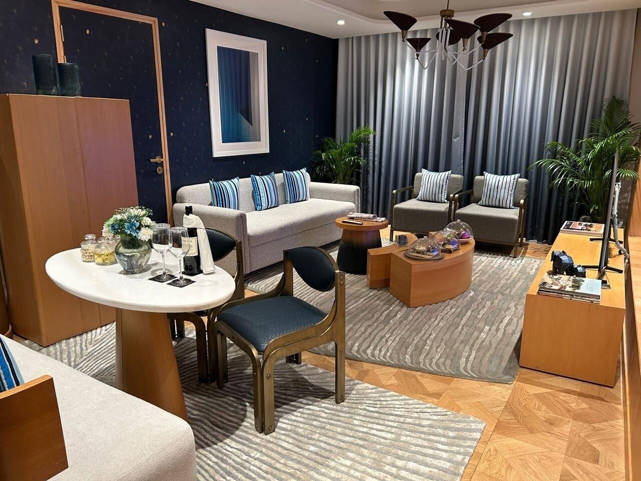A modern living room with a sofa set, a dining table, and a coffee table. There is a large window with curtains, a chandelier, and a potted plant.