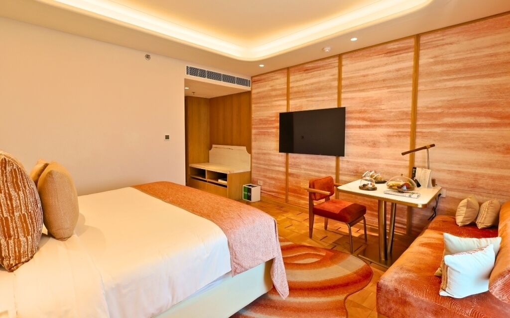 A hotel room with a bed, a desk, a TV, a sofa, and a small table with items on it.