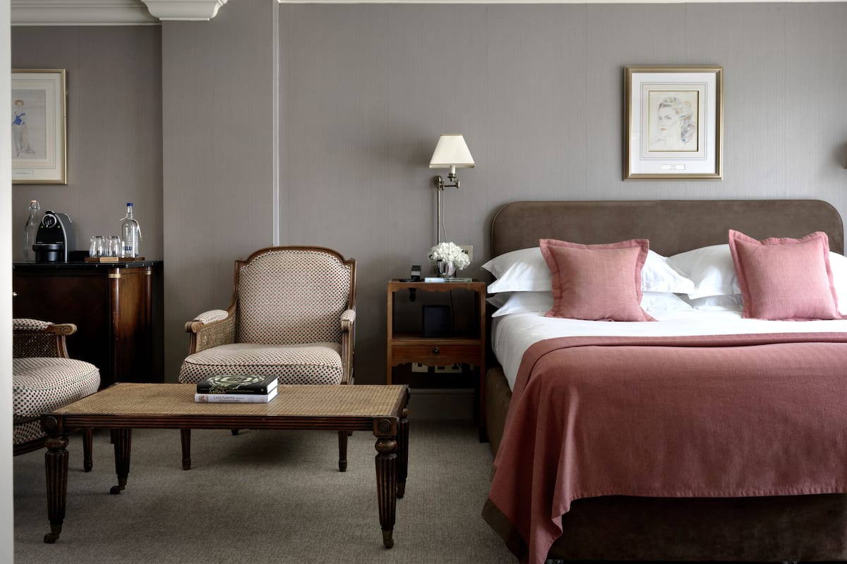 A hotel room with a large bed, a chair, a table, and a nightstand with a lamp.