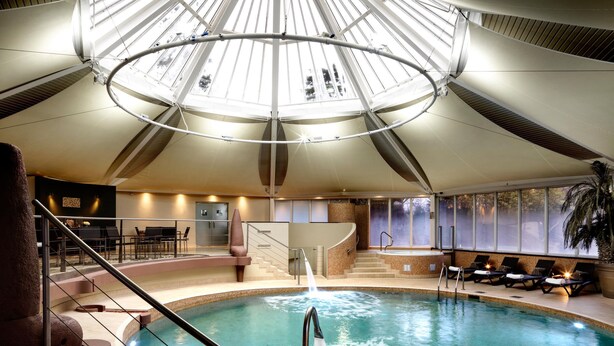 An indoor swimming pool with a glass ceiling, a seating area with chairs, and a staircase leading to the pool. - The Vineyard (Newbury)
