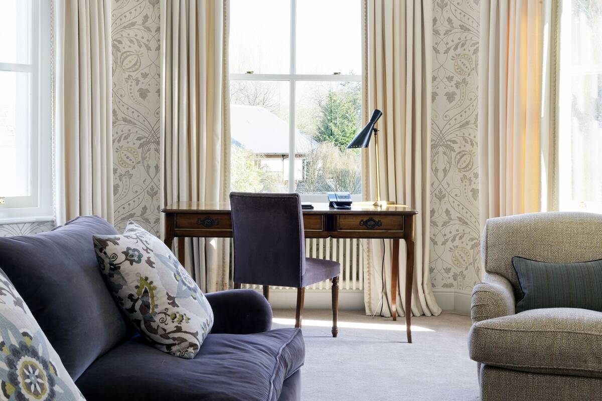 A room with a dark grey sofa, a patterned armchair, a wooden desk with a laptop, and a window with curtains.