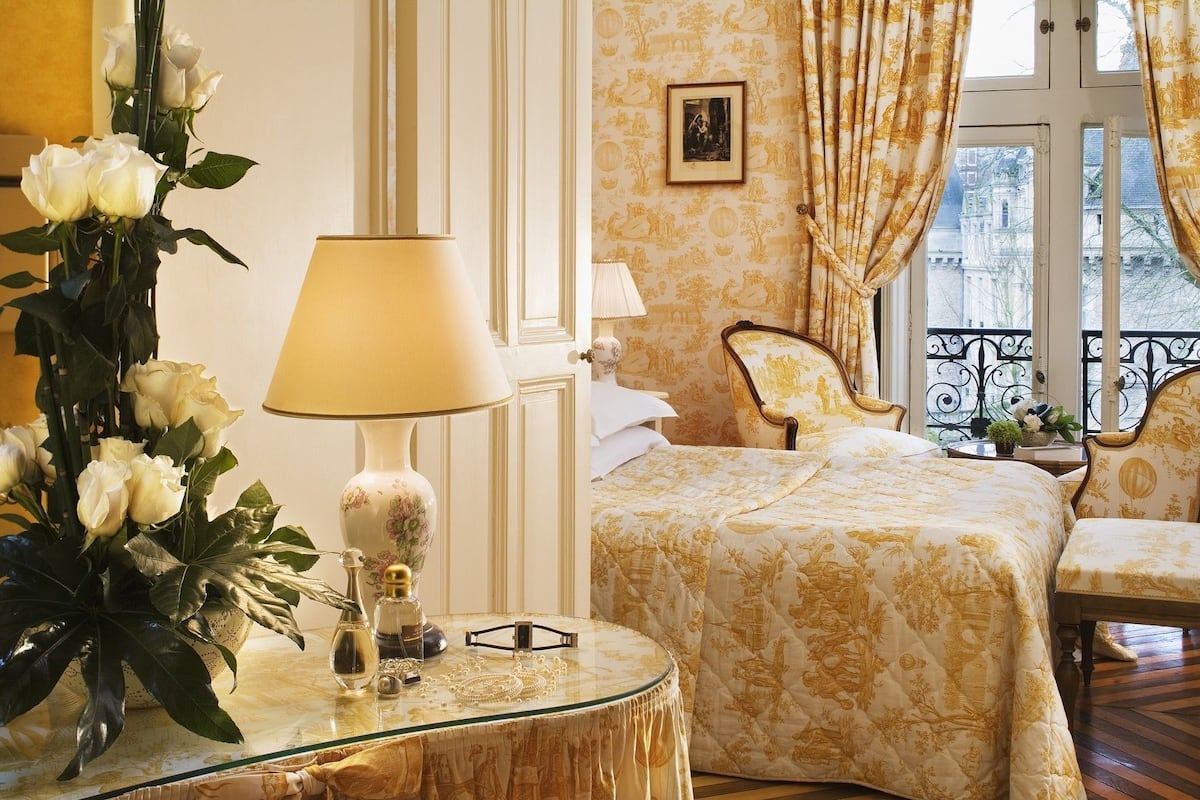 A hotel room with a bed, a chair, a table with a lamp, and a vase of flowers.