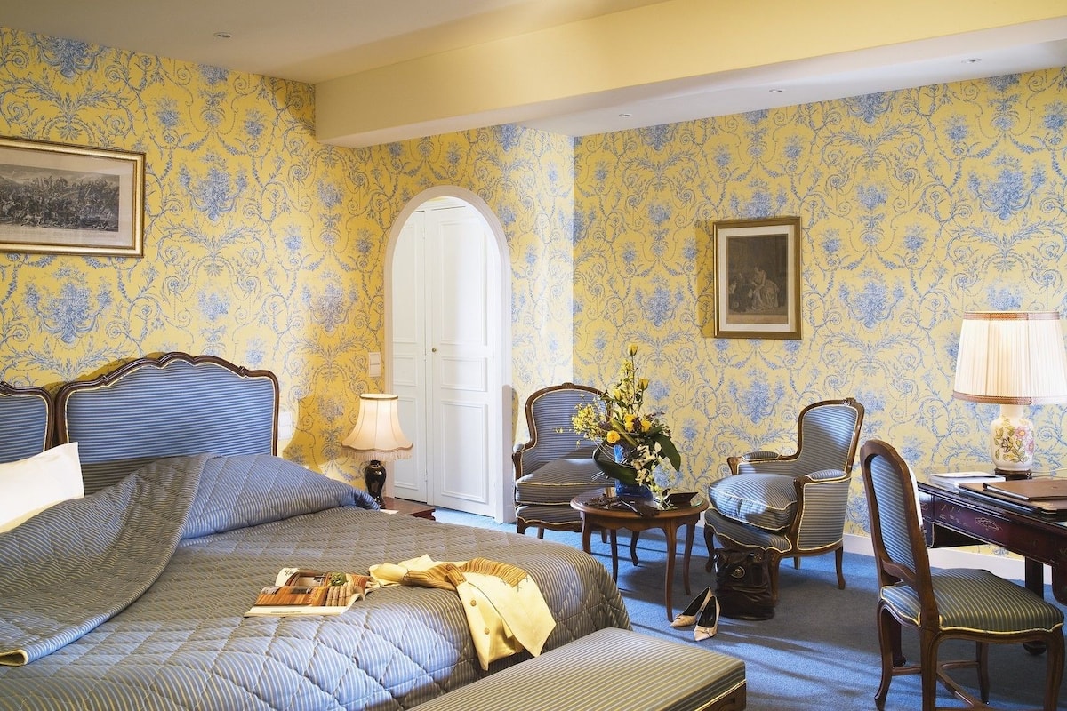 A room with a large bed, two armchairs, a small table with a vase of flowers, and a desk with a lamp.