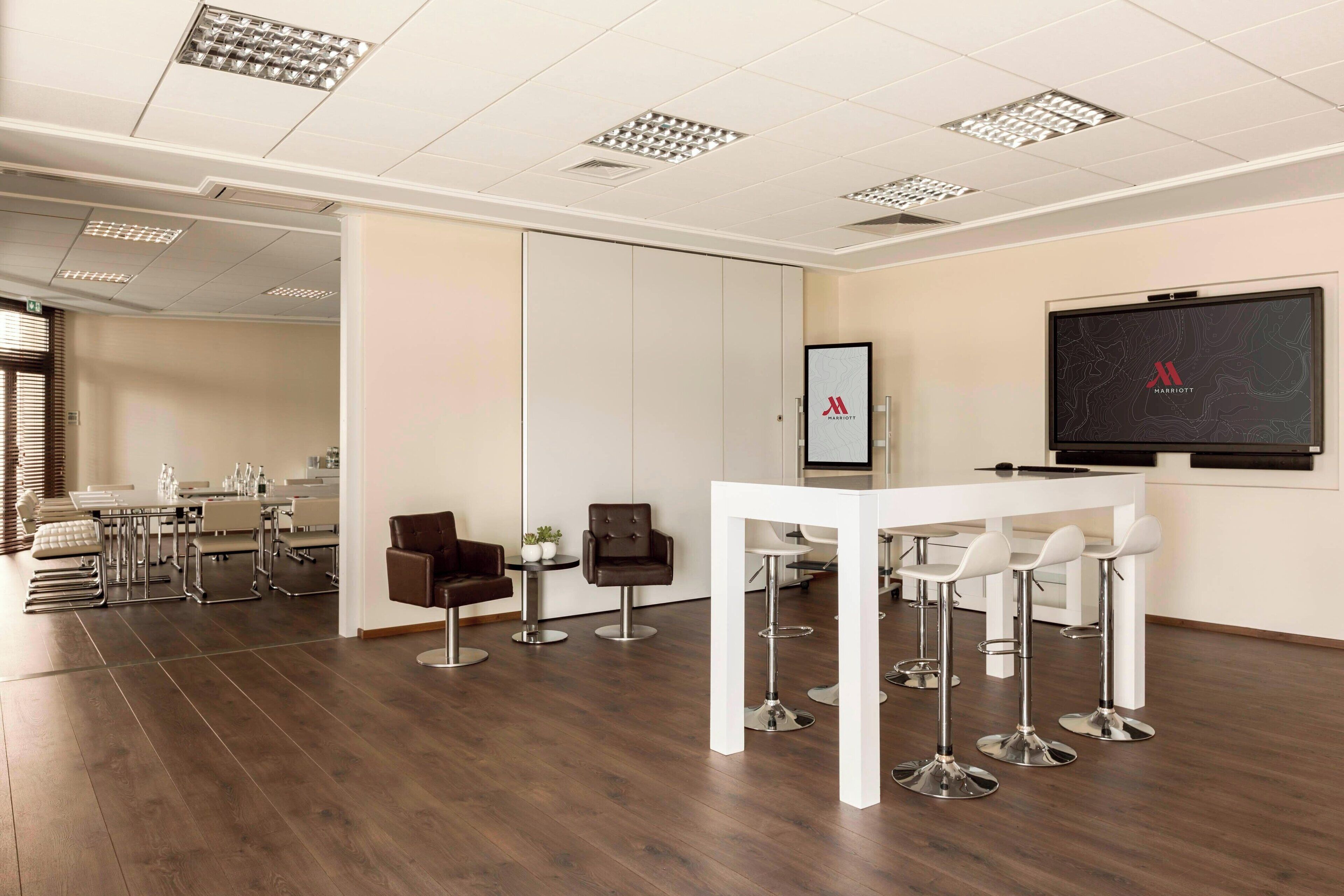 Una sala de conferencias de hotel moderna con una mesa blanca y taburetes de bar, una pantalla grande que muestra el logotipo de Marriott, y un área de comedor separada con una mesa y sillas blancas.
